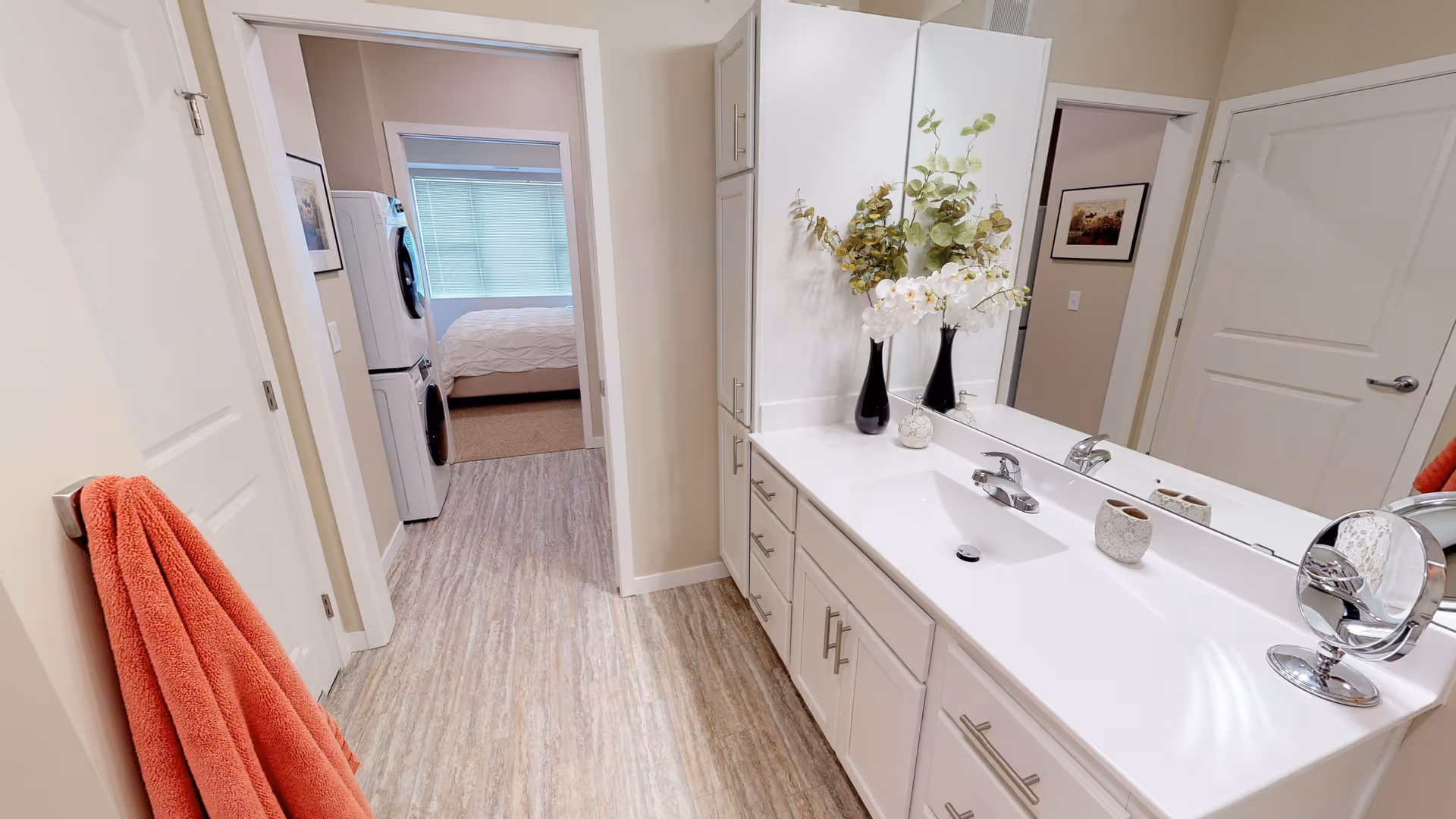 A bright bathroom with a long white countertop featuring a sink, a large mirror, and decorative vases with flowers. A coral towel hangs on a hook on the left wall. Through an open doorway, a stacked washer and dryer are visible, and beyond that, a bedroom with a bed and a window with blinds can be seen.