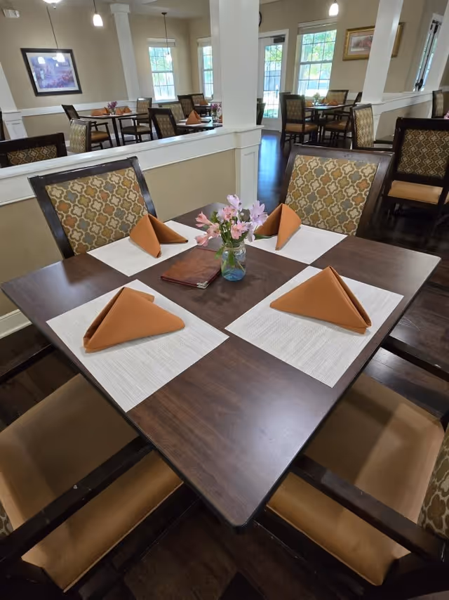 A dining table set for four with brown napkins folded in a triangular shape on white placemats. A small vase with pink flowers and a closed menu are placed in the center of the dark wooden table. The room has patterned cushioned chairs, beige walls, large windows letting in natural light, and several other similarly arranged tables in the background.