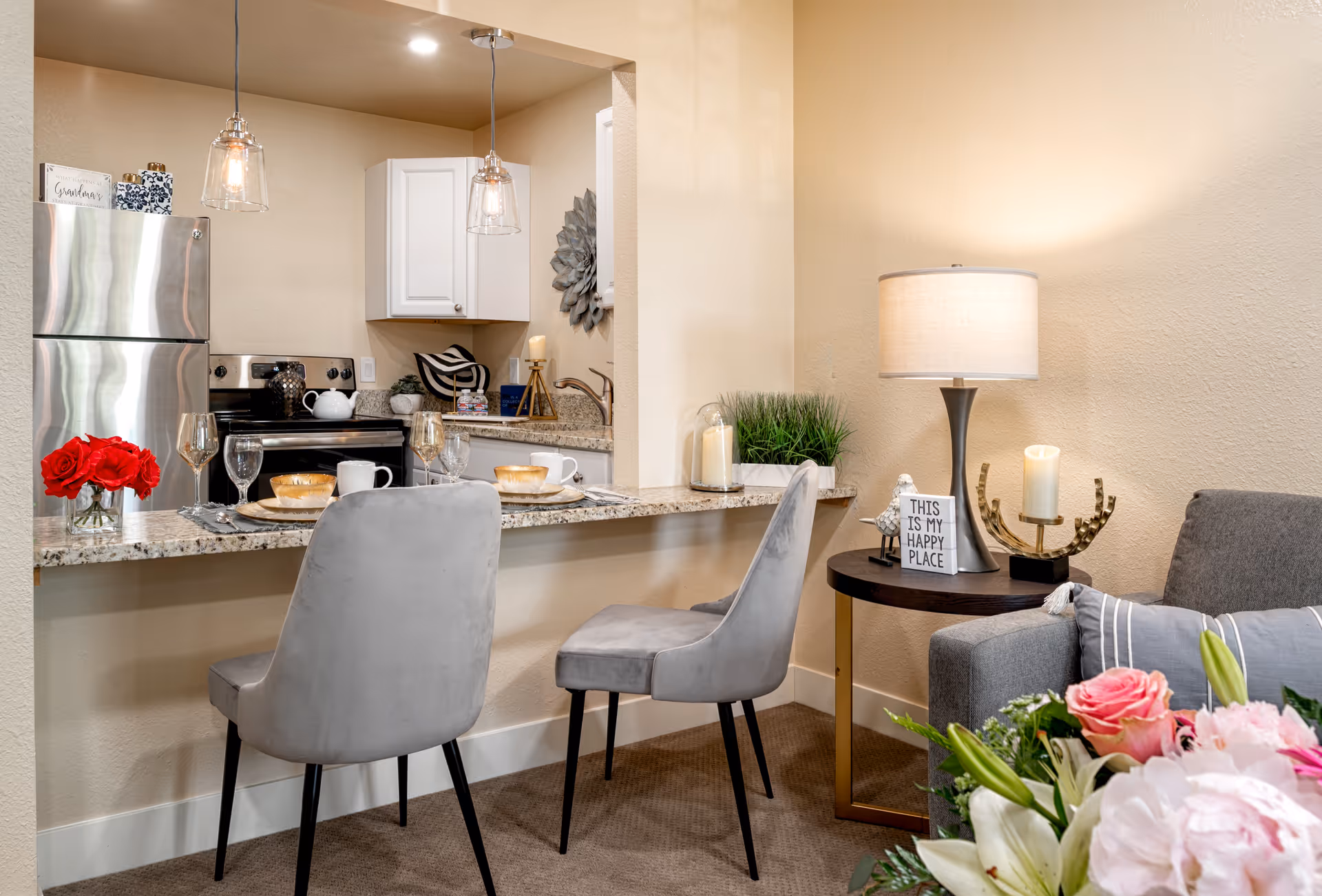 A cozy interior space featuring a small kitchen with stainless steel refrigerator, stove, and white cabinets. In front of the kitchen counter are two gray upholstered chairs set for dining with plates, glasses, and a vase of red roses. To the right is a living area with a gray sofa, a side table with a lamp, decorative candles, and a sign that reads 'This is my happy place.' A bouquet of flowers is visible in the foreground.