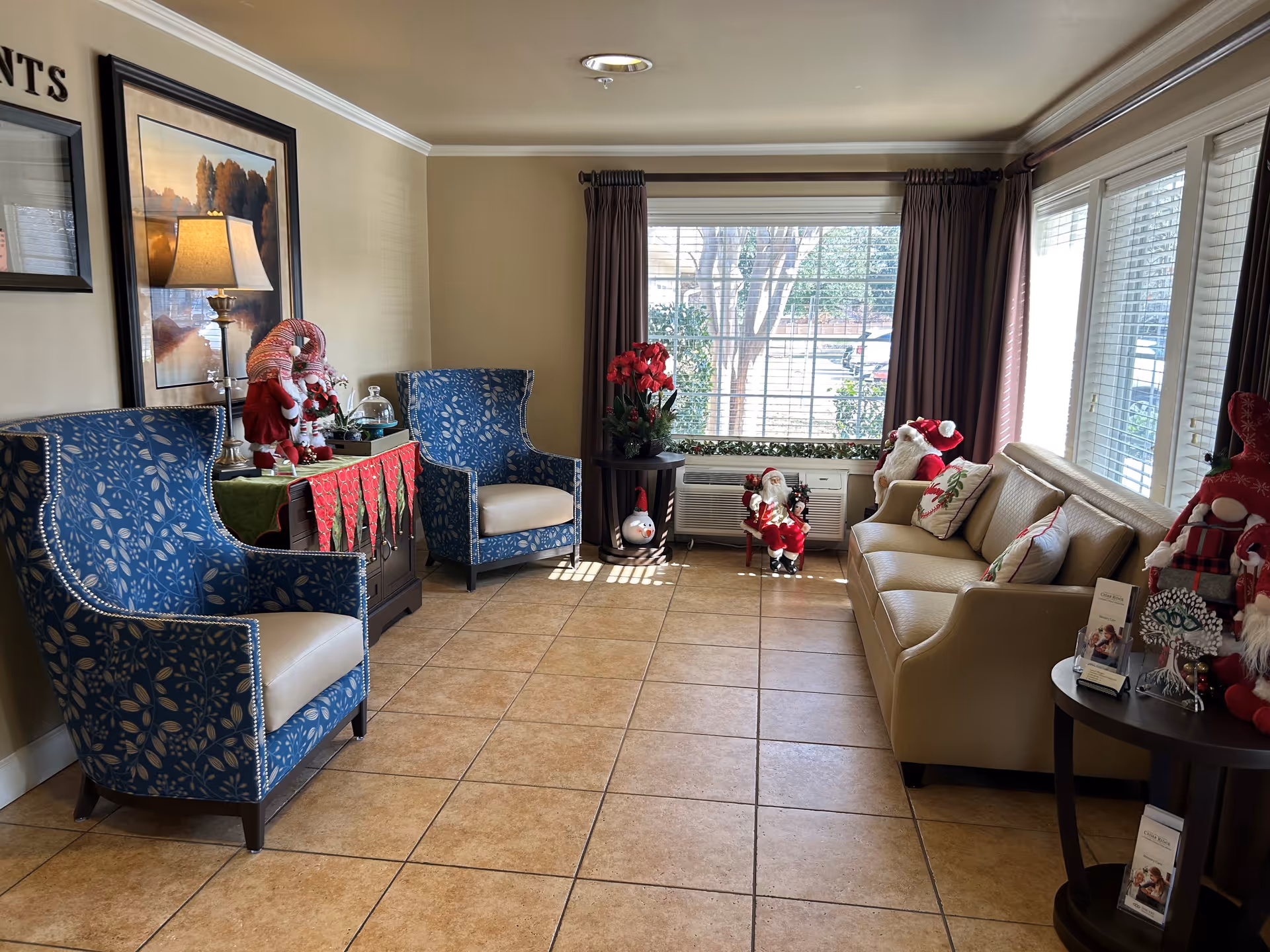 A cozy living room area with two blue patterned armchairs, a beige sofa with decorative pillows, and a side table. The room is decorated with Christmas-themed items including Santa figurines and red flowers. Large windows with brown curtains allow natural light to fill the space.