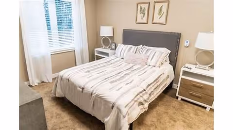 A bedroom with a neatly made bed featuring striped bedding and multiple pillows. The bed has a dark upholstered headboard. On each side of the bed, there are white nightstands with drawers, each holding a modern lamp with a circular base. Two framed botanical prints hang on the beige wall above the headboard. A window with white blinds and sheer curtains is visible on the left side of the room.