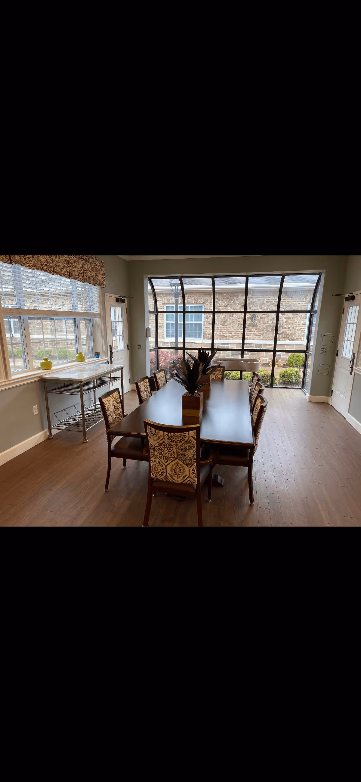 A dining room with a long rectangular wooden table surrounded by eight upholstered chairs with patterned backs. The room has large windows on two sides, allowing natural light to fill the space. There is a decorative centerpiece on the table and a small metal rack with a white countertop against one wall. The floor is wooden, and the walls are painted light gray.