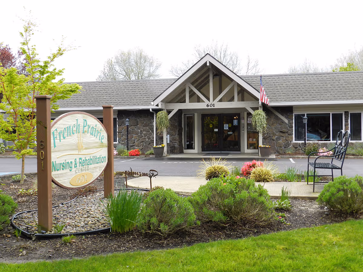 Front exterior view of French Prairie Nursing & Rehabilitation Center building with a peaked roof entrance, stone facade, American flag, and landscaped garden with bushes and flowers. A wooden sign with the facility name and address 601 is visible near the sidewalk.