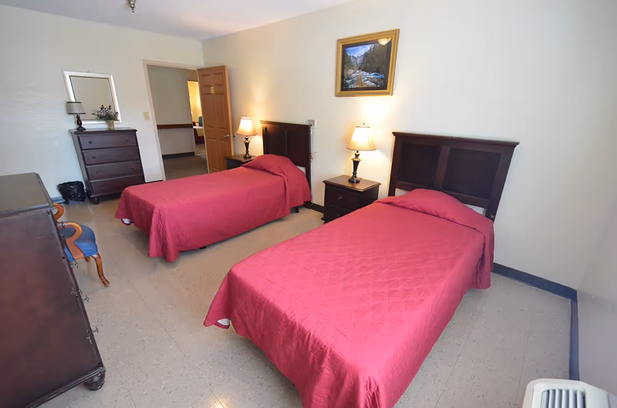 A bedroom with two single beds covered in red bedspreads, each with a dark wooden headboard. Between the beds is a nightstand with a lamp. On the left side of the room, there is a dresser with a mirror and a small decorative plant. The room has a tiled floor and light-colored walls, with a framed landscape painting hanging above the nightstand. An open door leads to another room.