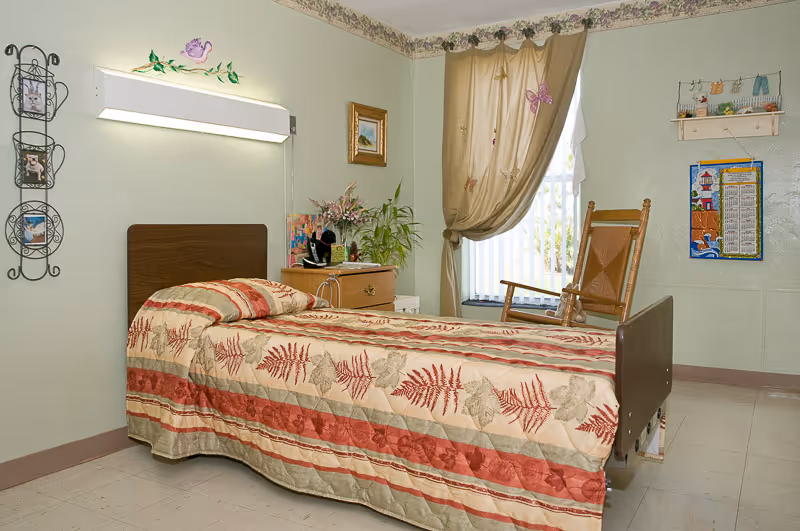 A single bed with a patterned bedspread featuring red and beige leaves in a room with light green walls. Next to the bed is a wooden nightstand with a plant and other small items. A wooden rocking chair is placed near a window with beige curtains decorated with butterflies. The room has decorative wall hangings and a floral border near the ceiling.