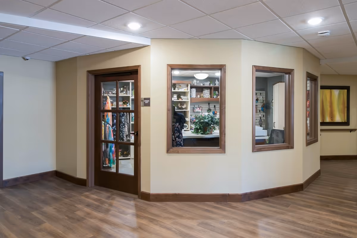 Interior hallway of a senior living facility with wood flooring and beige walls. There is a wooden door with glass panels labeled 'BOUTIQUE GIFTS' leading to a small gift shop. The shop has shelves displaying various items including clothing and decorative pieces. There are also two large windows looking into the gift shop from the hallway.