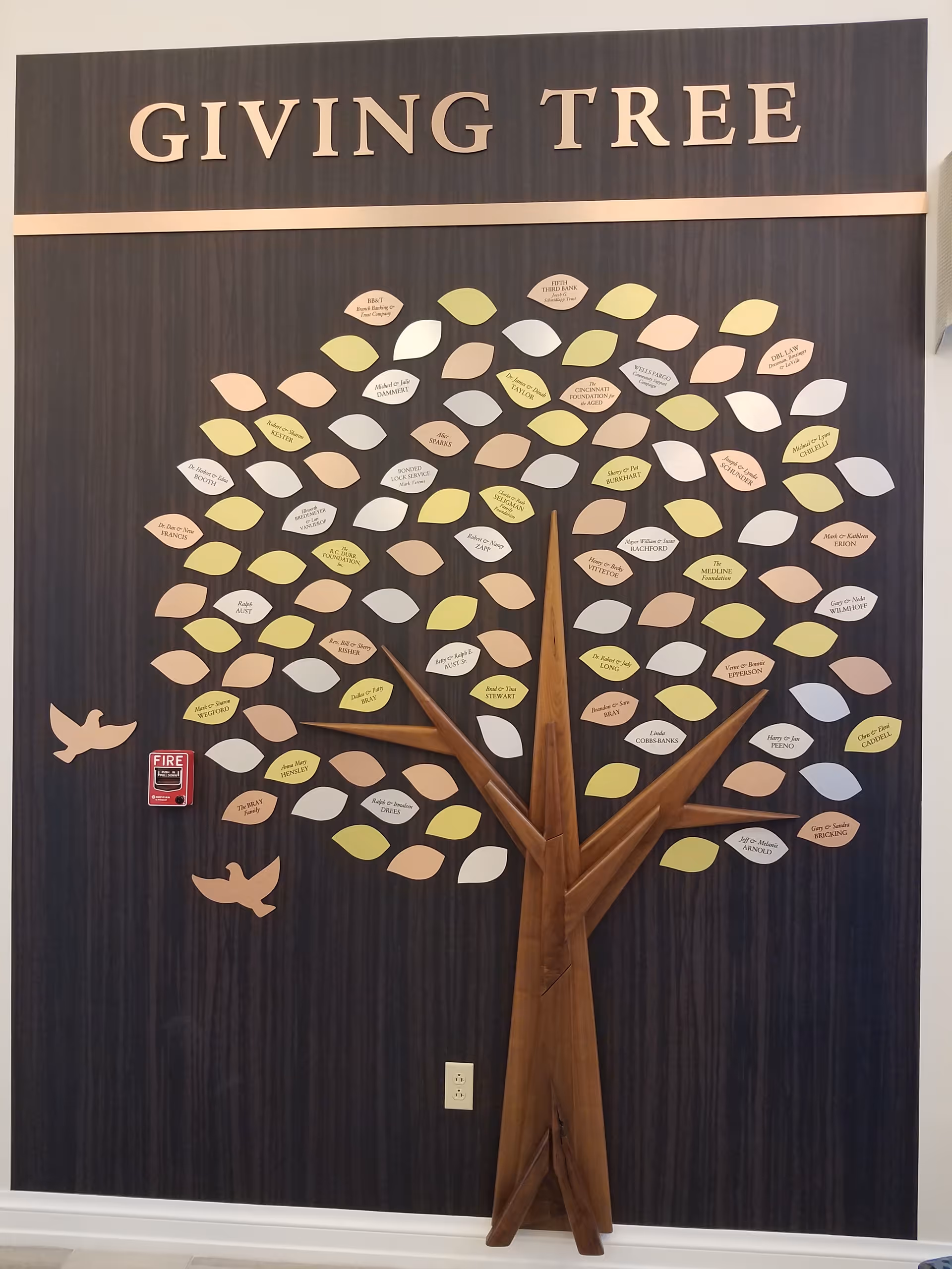 Decorative 'Giving Tree' wall display featuring a wooden tree trunk and many colored leaf plaques on a dark paneled interior wall.