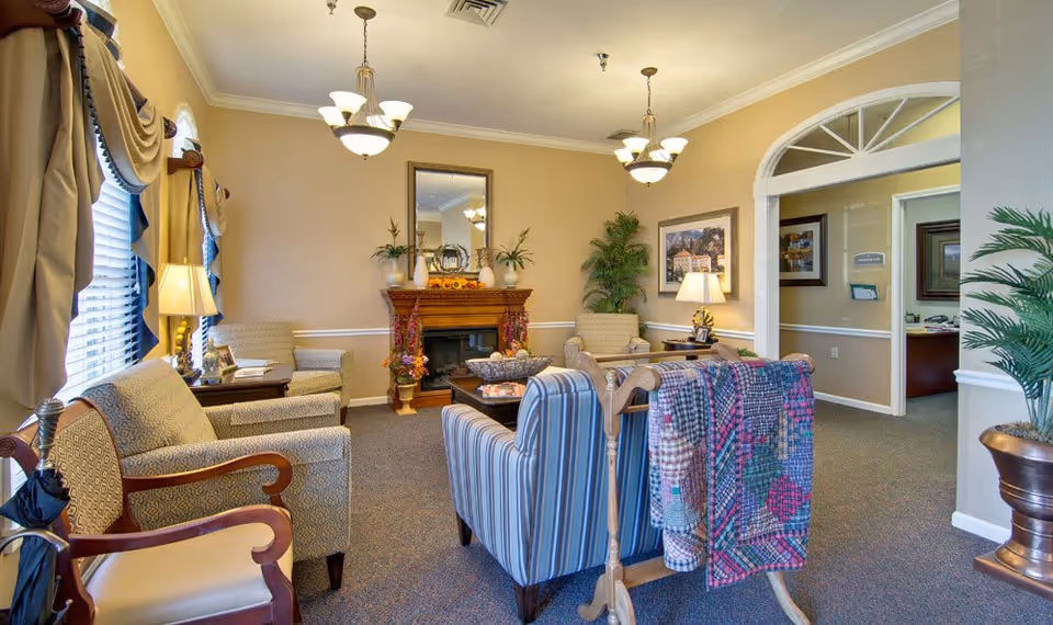 A cozy senior living facility common area with comfortable armchairs arranged around a wooden coffee table. The room features a decorative fireplace with plants and a mirror above it, warm beige walls, two hanging light fixtures, and large windows with draped curtains. A colorful quilt is draped over a wooden stand, and there are framed pictures and lamps on side tables. An open doorway leads to an office area in the background.