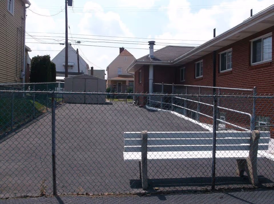 Outdoor paved area enclosed by a chain-link fence with a white bench inside. There is a red brick building on the right side and a small shed at the far end. Residential houses and utility poles are visible in the background under a partly cloudy sky.