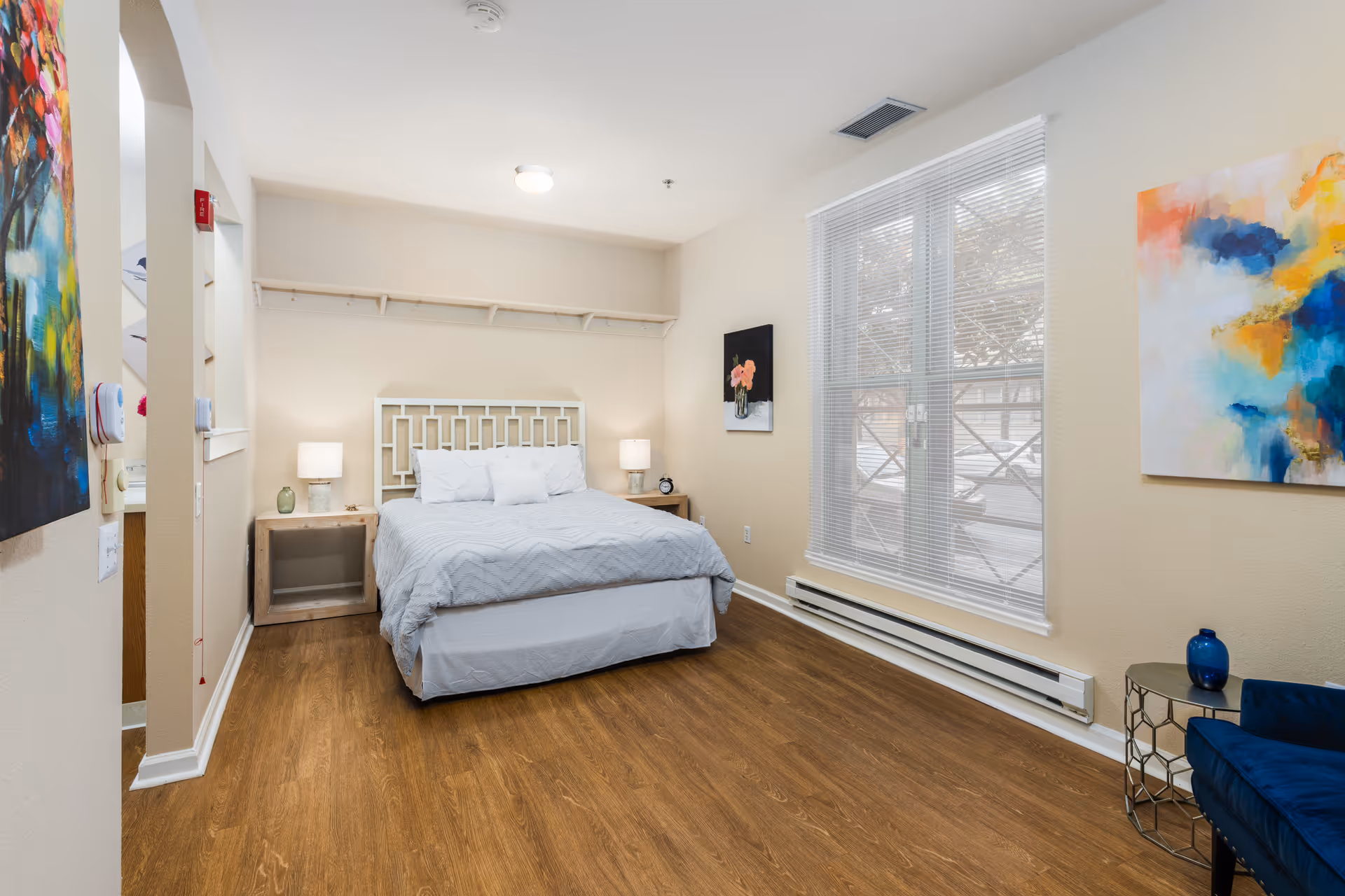 Bright furnished bedroom with a bed, nightstands, large window, and colorful wall art.