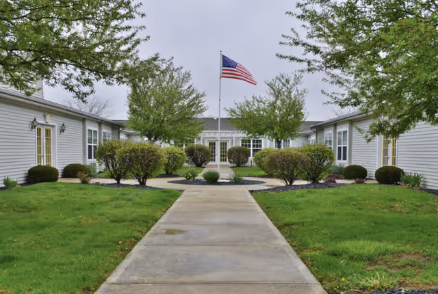 A courtyard area at Trustwell Living at Oakley Place featuring a concrete walkway leading to a building entrance. The courtyard is landscaped with green grass, trimmed bushes, and several trees. An American flag is flying on a flagpole in the center of the courtyard.