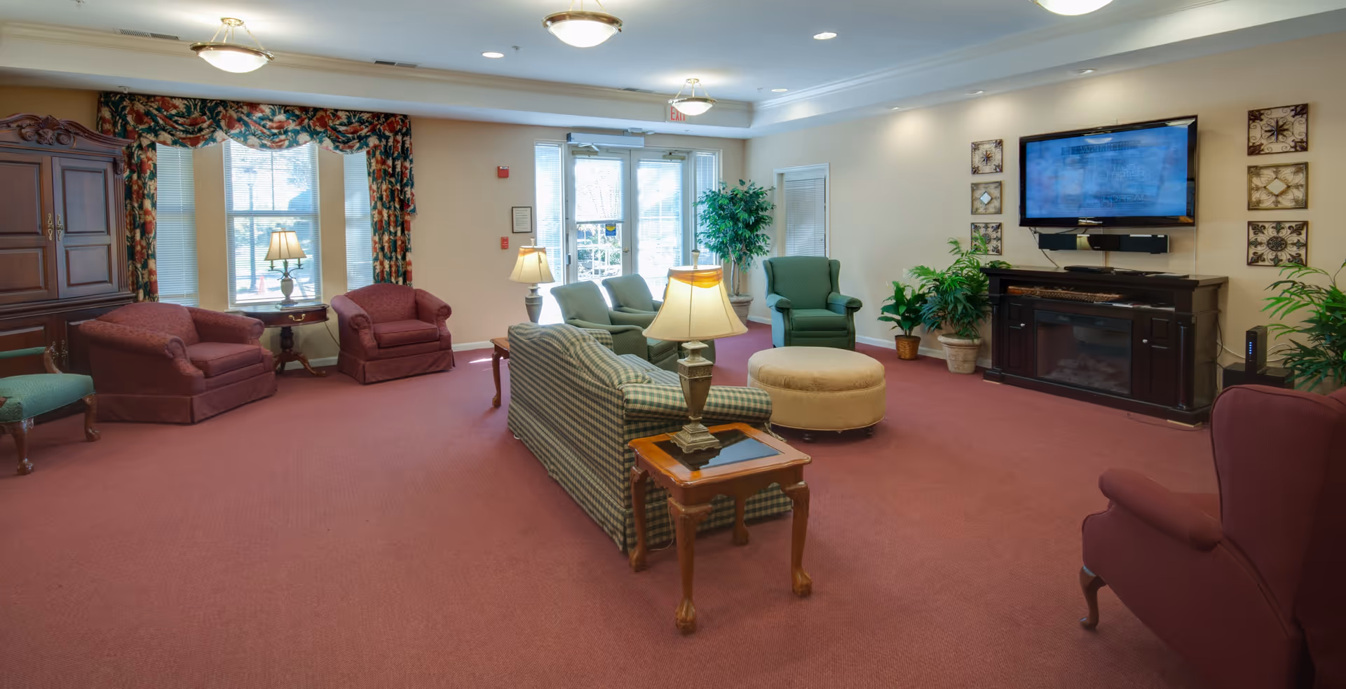 A spacious senior living facility common area with various upholstered chairs and sofas arranged around a central ottoman. The room features a large flat-screen TV mounted above a dark wooden electric fireplace, several potted plants, and large windows with floral curtains allowing natural light to enter. The carpet is a muted red color, and the walls are painted a soft beige.