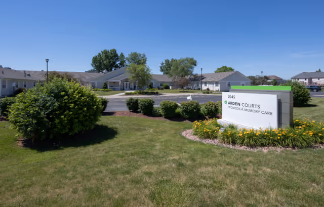 Exterior view of Arden Courts - ProMedica Memory Care Community in South Holland, showing a single-story building with a parking lot, green lawn, bushes, and a sign with the facility's name and address under a clear blue sky.
