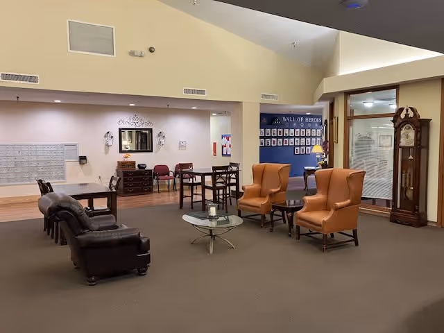 Community lounge in a retirement facility with leather sofas and armchairs arranged around coffee tables, a 'Wall of Heroes' display, and a grandfather clock.