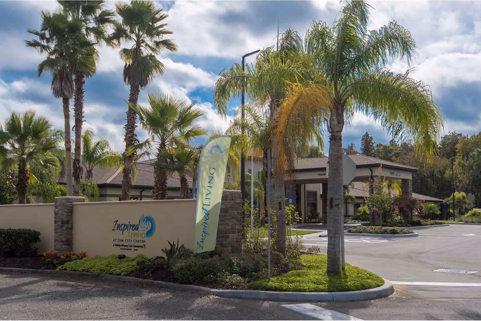 Entrance to Inspired Living Sun City Center facility with palm trees, a stone and beige wall with the facility name and logo, a flag with Inspired Living text, and a driveway leading to the building under a partly cloudy sky.