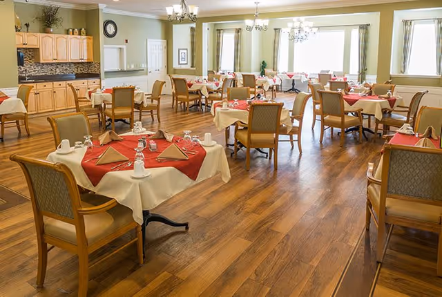 Dining room with multiple tables set with red napkins and wooden chairs on a wood floor under chandeliers and large windows.