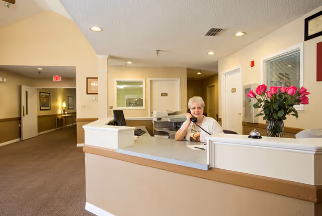 Reception area of Bradfield Terrace senior living facility with a smiling woman sitting behind the front desk, talking on the phone. The desk has a computer, a bell, and a vase with pink roses. The background shows a hallway with doors and framed pictures on the walls.