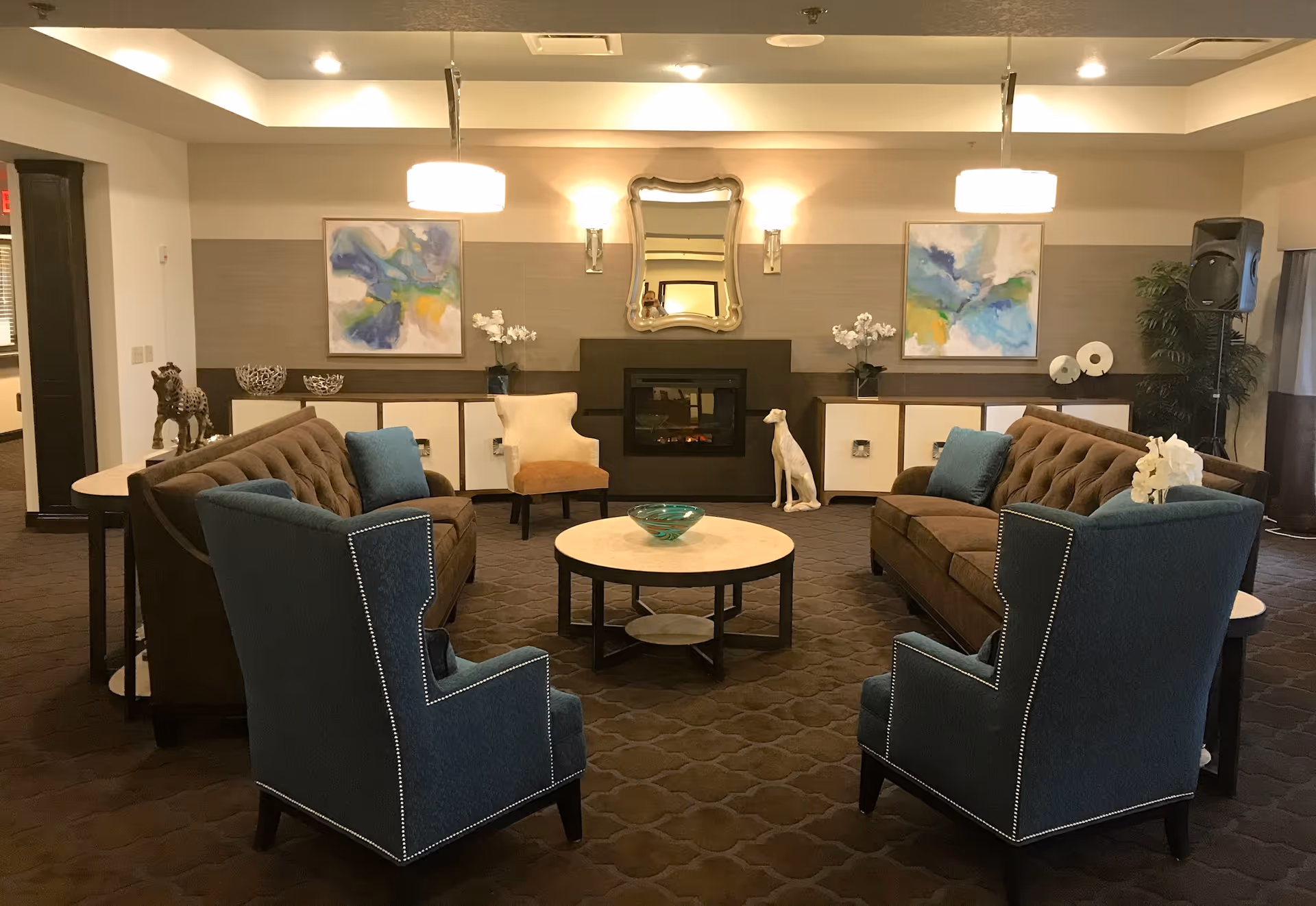 A cozy living room area with two brown tufted sofas facing each other, two blue armchairs with nailhead trim, a round coffee table with a decorative bowl, and a fireplace with a decorative mirror above it. The room is decorated with abstract paintings, white orchids, and a dog statue, with warm lighting from ceiling fixtures and wall sconces.