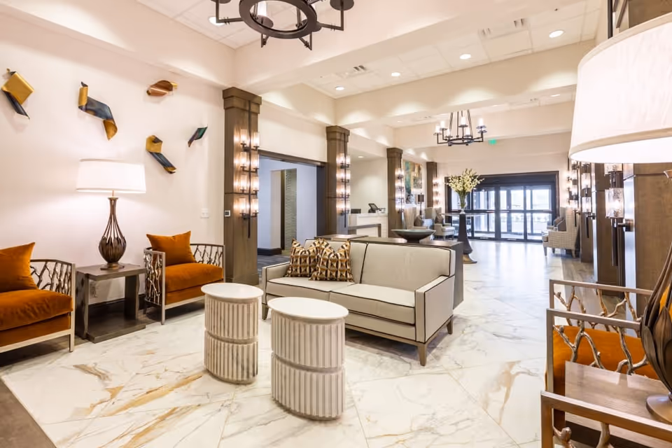 A spacious and well-lit lobby area with modern furnishings including a beige sofa with patterned pillows, two orange armchairs, two round white side tables, and decorative wall art. The floor is covered with large marble tiles, and there are multiple light fixtures on the walls and ceiling. In the background, there is a reception desk and glass entrance doors.