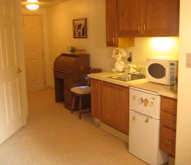 A small kitchen area with wooden cabinets, a white mini refrigerator with magnets, a microwave on the countertop, and a sink. To the left, there is a wooden roll-top desk with a chair and a framed picture on the wall above it. The floor is covered with light-colored linoleum, and there is a door at the end of the hallway.