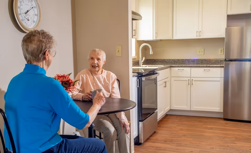 Two older women sit and converse at a small round table in a kitchen/dining nook with white cabinets and stainless steel appliances.