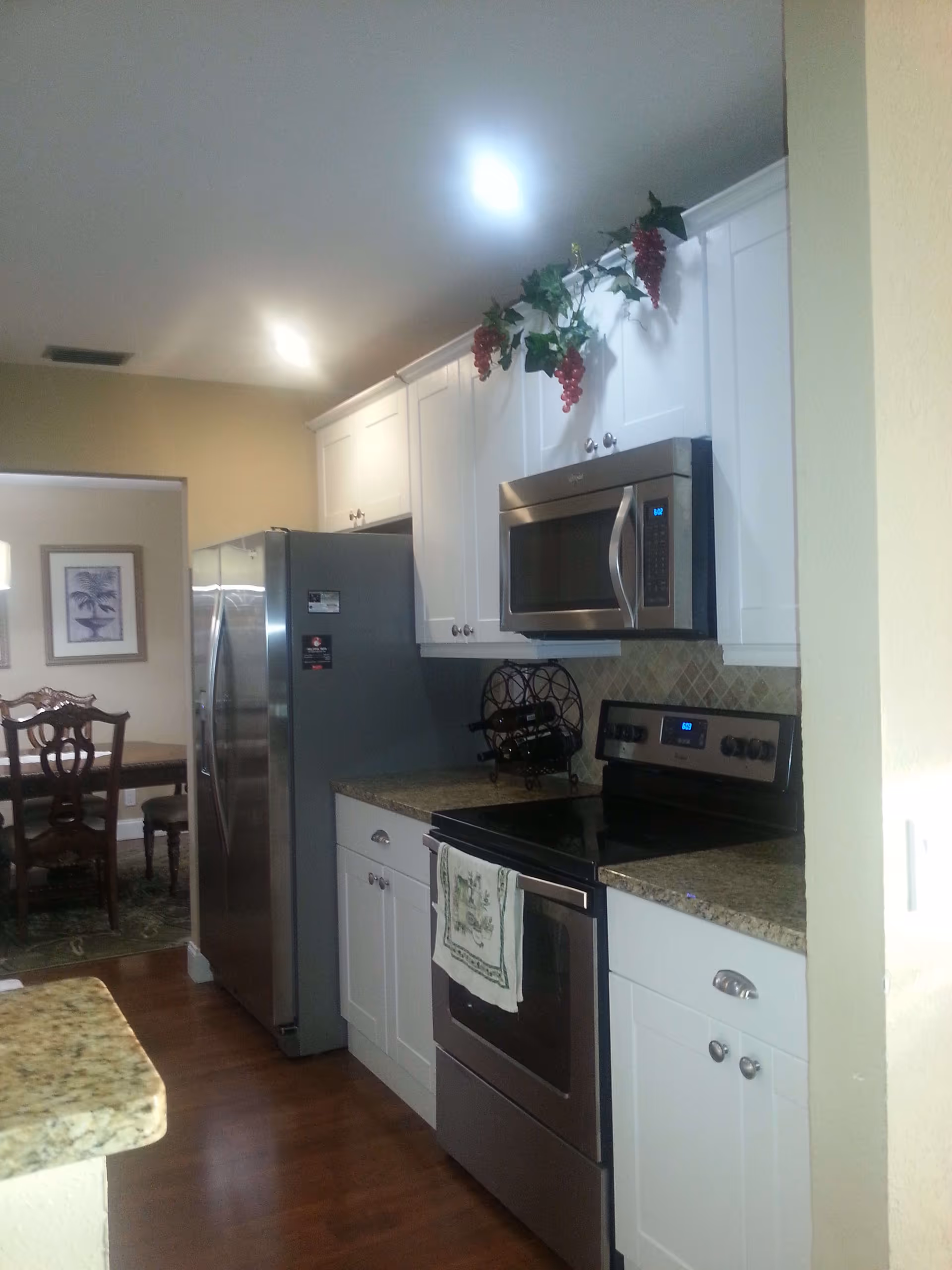 Modern kitchen with white cabinets, granite countertops, stainless steel refrigerator, microwave, and stove. A decorative grapevine is placed above the cabinets. In the background, a dining area with a wooden table and chairs is visible.