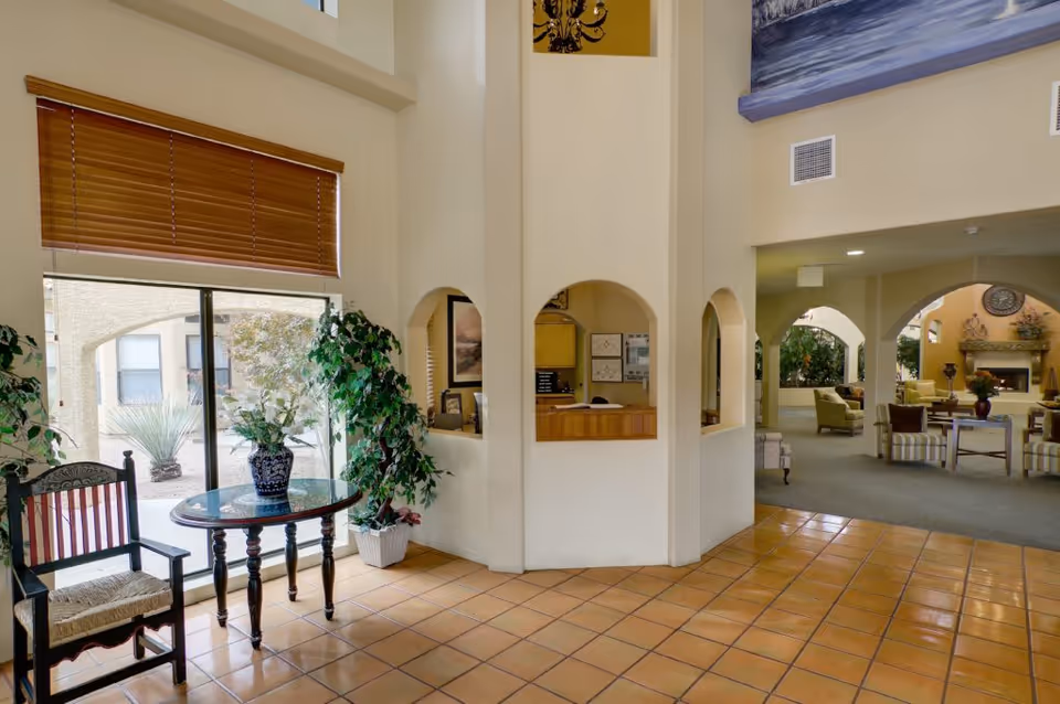 Interior view of a senior living facility lobby area with terracotta tile flooring, a round wooden table with a floral vase, a wooden chair, and large windows with wooden blinds. The space includes a reception desk with arched openings and a seating area with armchairs and a fireplace in the background.