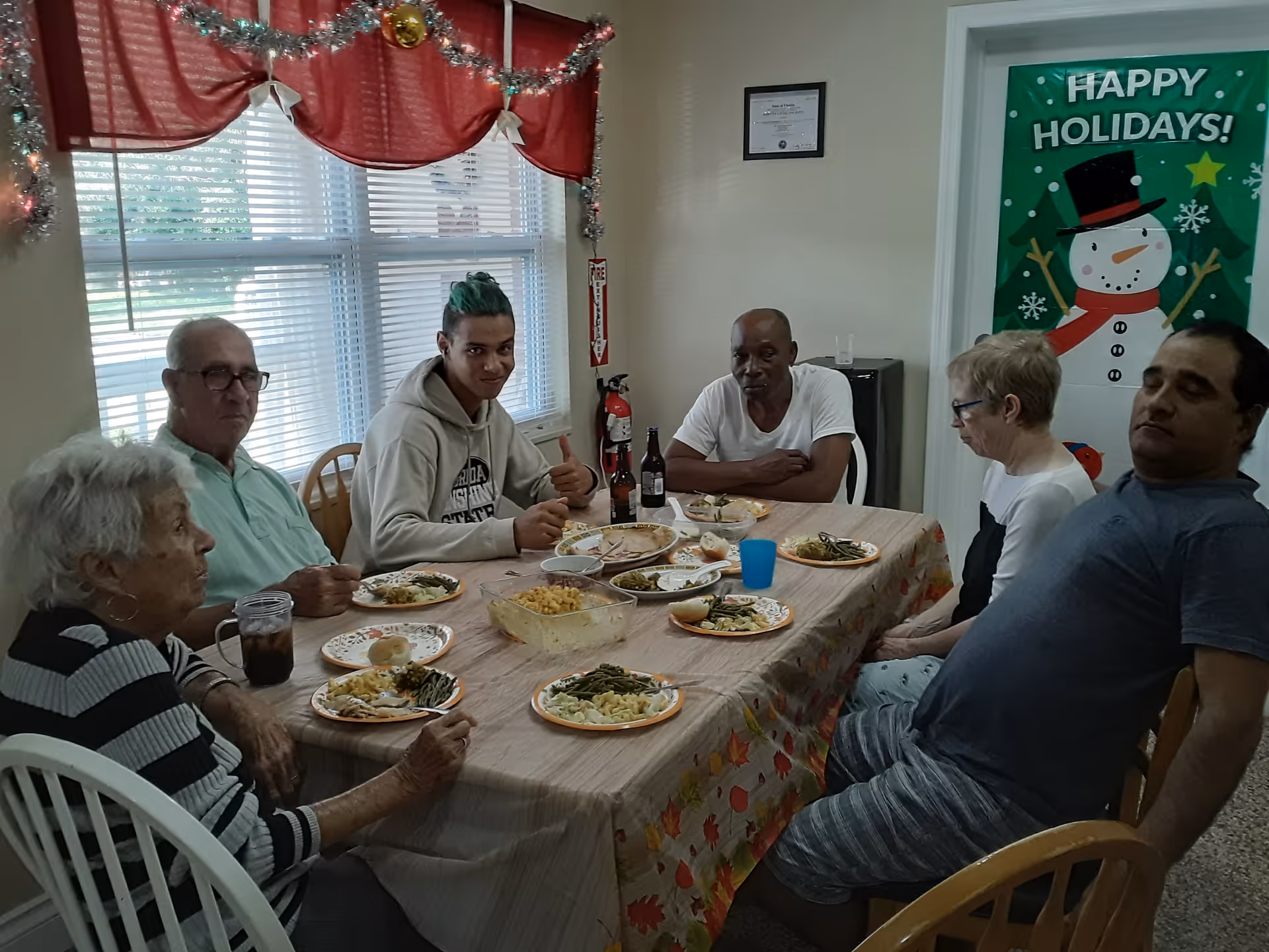Several people sit around a decorated table eating a meal in a dining room with a 'Happy Holidays' poster on the wall.