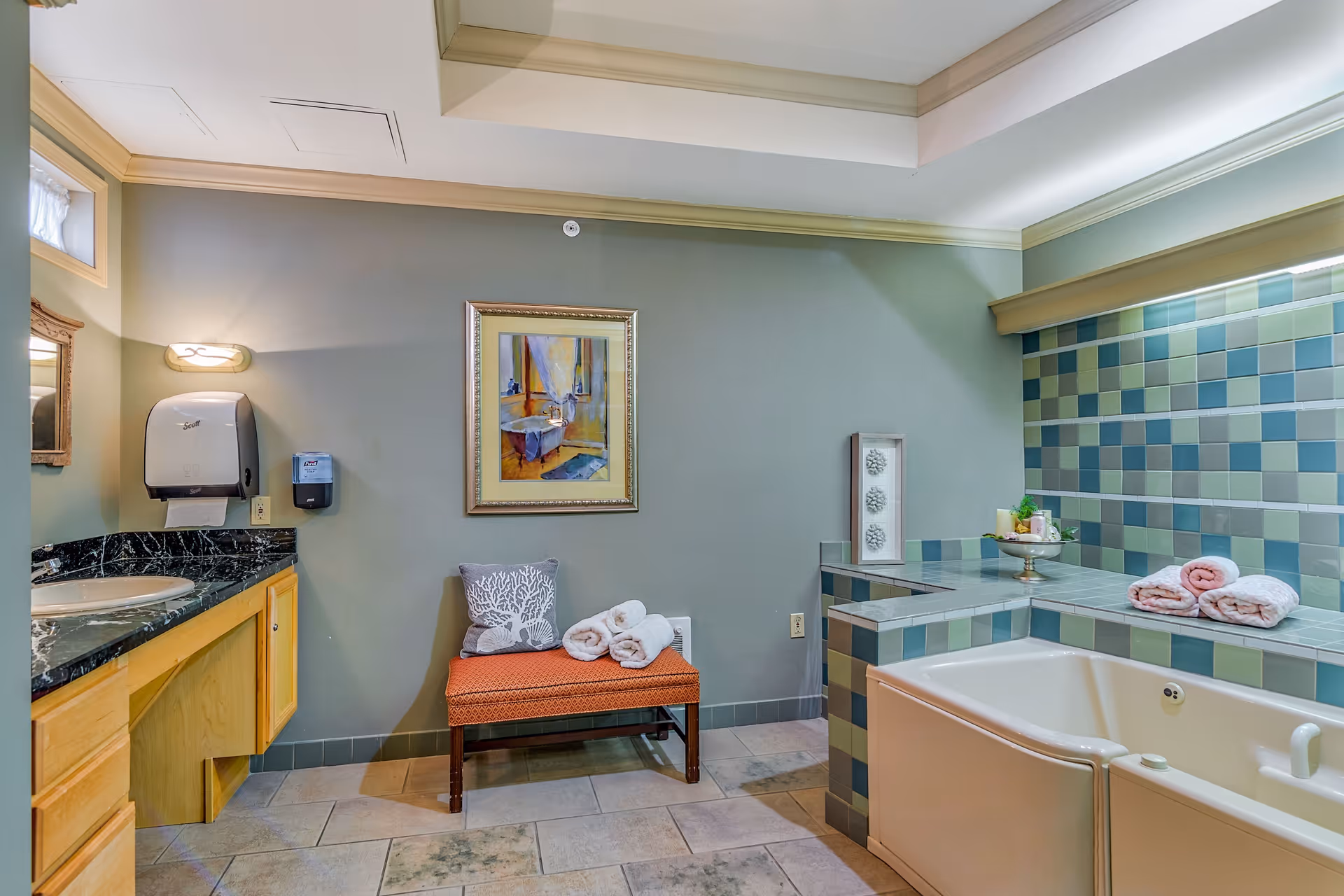 A bathroom with a large bathtub surrounded by blue and green square tiles, a wooden vanity with a black marble countertop and sink, a wall-mounted paper towel dispenser and soap dispenser, a small bench with a decorative pillow and rolled white towels, and a framed painting on the wall.