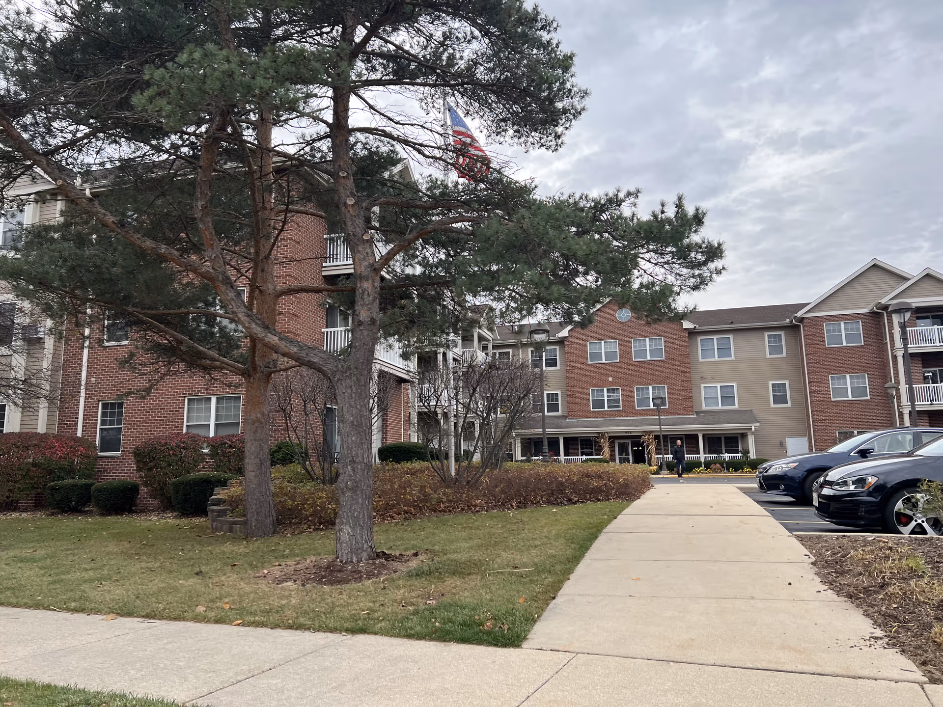 Exterior view of a multi-story senior living facility building with brick and beige siding, surrounded by trees, shrubs, and a lawn. A concrete walkway leads to the entrance, and several parked cars are visible on the right. An American flag is flying above the building under a cloudy sky.