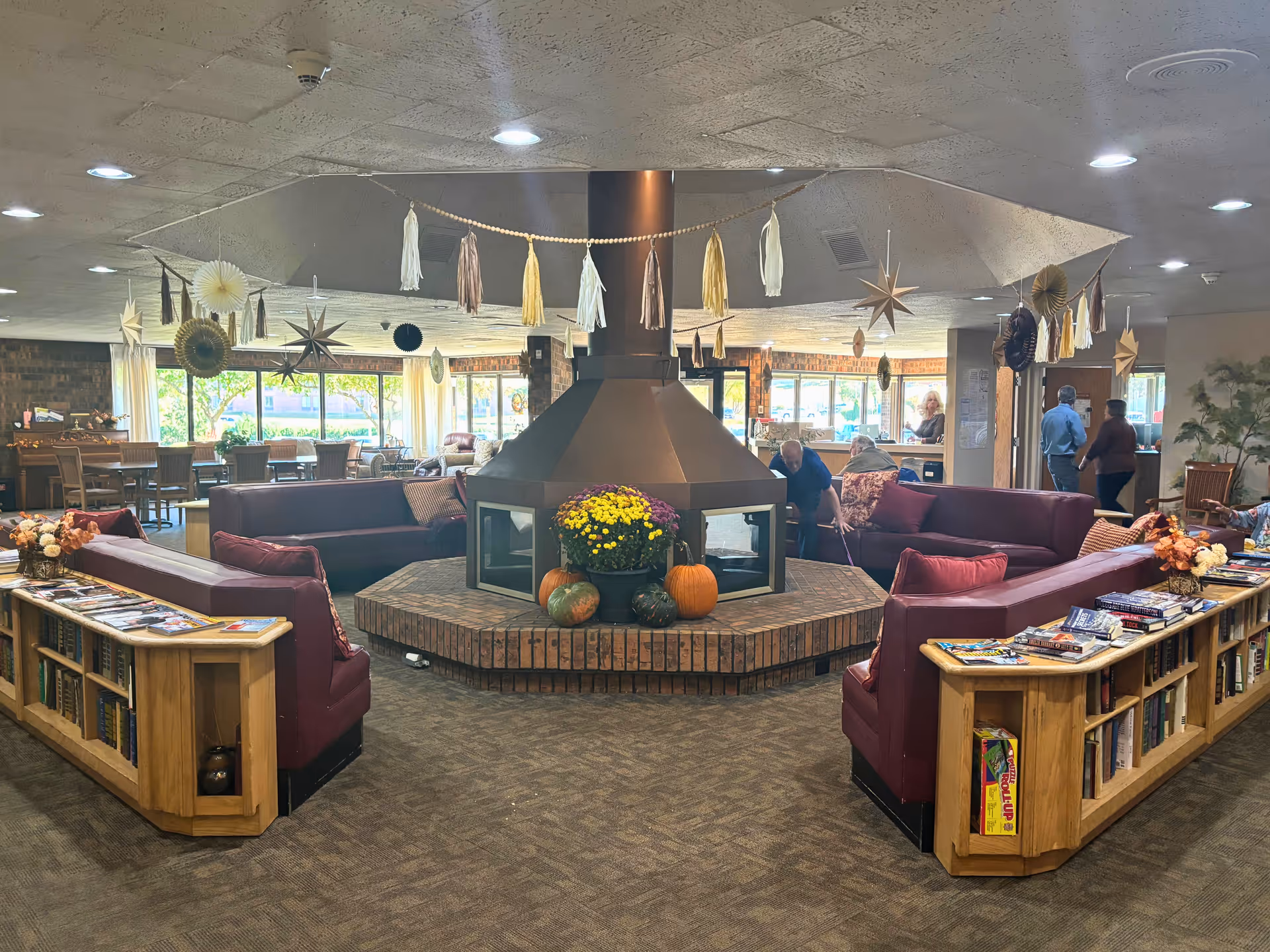 Communal lounge area with a central hexagonal fireplace, maroon sofas, bookshelves, and seasonal pumpkins and hanging decorations.