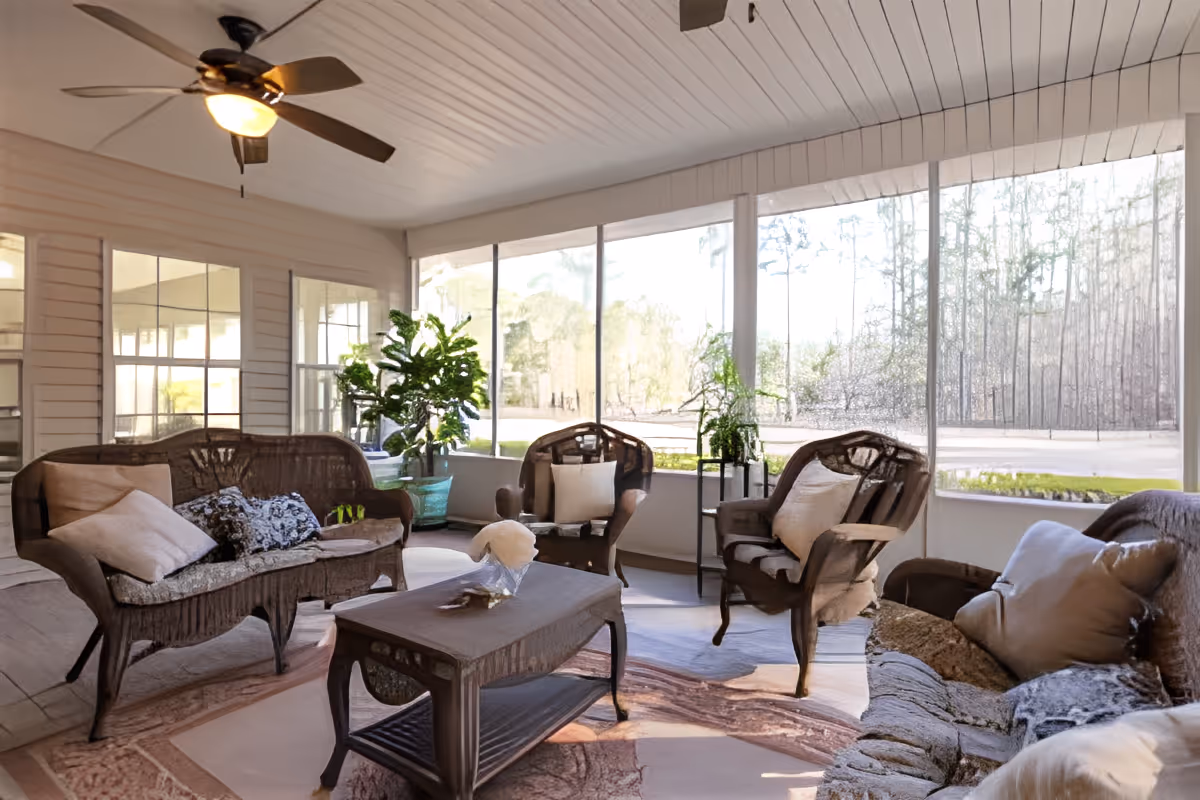 A bright and cozy sunroom with wicker furniture including two armchairs, a loveseat, and a coffee table. The room has large windows allowing natural light to fill the space and offers a view of trees outside. There are several potted plants adding greenery, a ceiling fan with a light fixture, and a patterned rug on the floor.