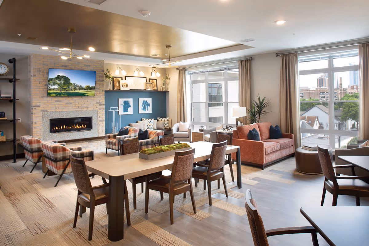 A bright and modern living room area in a senior living facility with large windows showing an urban view. The room features a brick fireplace with a mounted TV above it, several comfortable armchairs and sofas in warm tones, a long wooden table with chairs, decorative plants, and framed artwork on the walls.