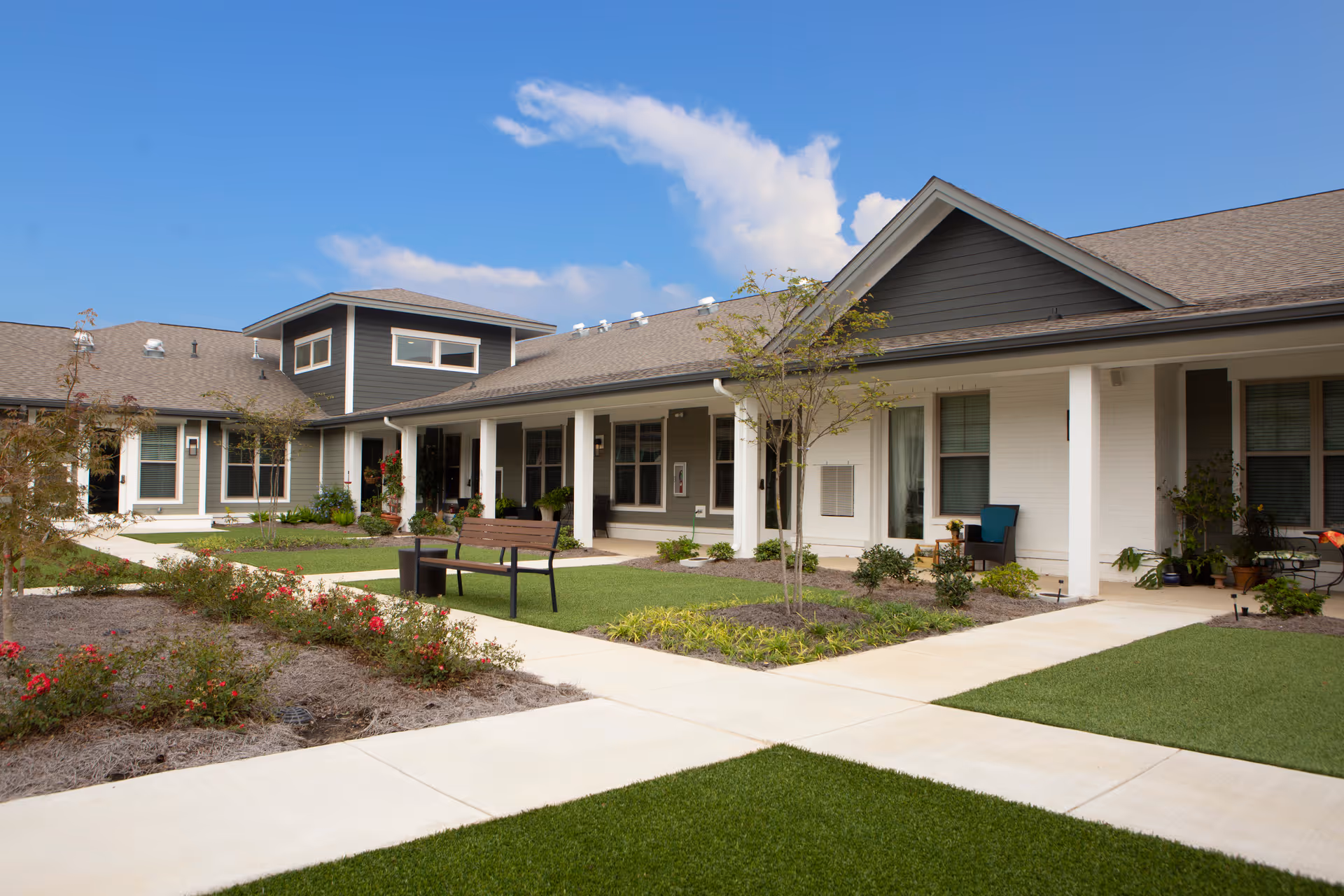 Courtyard and exterior facade of a single-story senior living building with walkways, landscaping, benches, and covered porches under a blue sky.