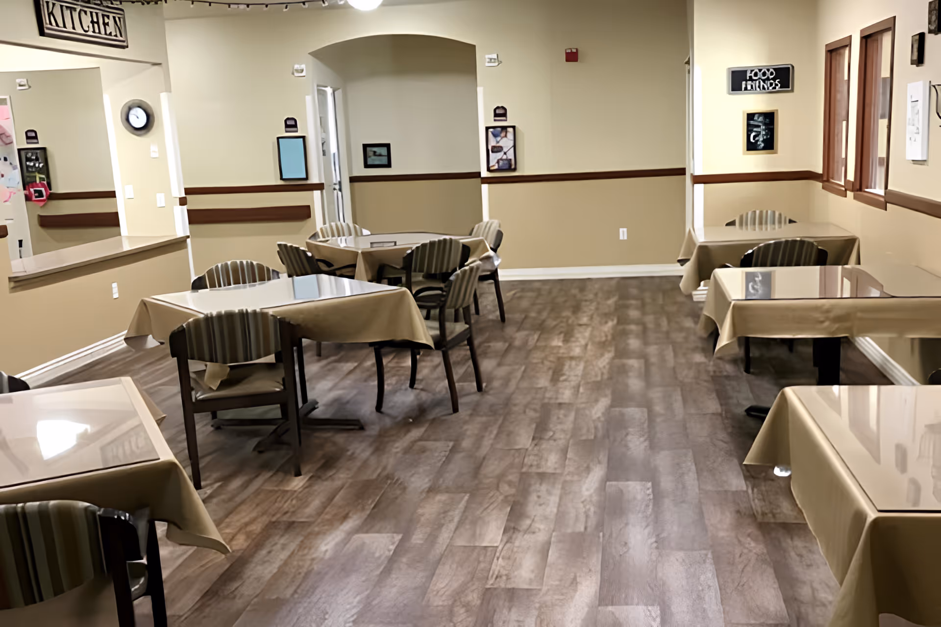 A dining room with several tables covered with beige tablecloths and surrounded by chairs. The room has wood flooring, beige walls with a wooden chair rail, and several windows on the right side. There is a sign above an open kitchen pass-through window that reads 'KITCHEN' and another sign on the right wall that says 'FOOD FRIENDS'.