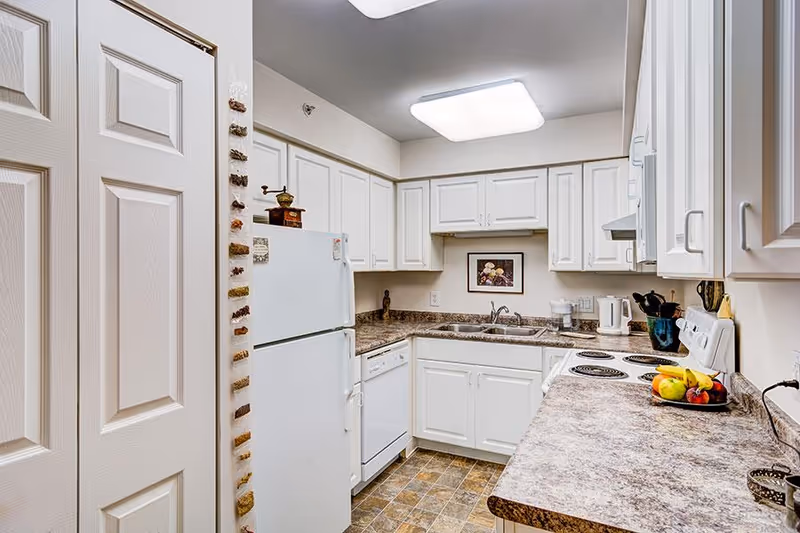 A compact kitchen with white cabinets and appliances including a refrigerator, dishwasher, stove, and microwave. The countertops are a brown and beige marble pattern, and there is a double sink under a framed floral picture on the wall. The floor has a tile pattern in shades of brown and beige. Various kitchen utensils and a bowl of fruit are on the counter.