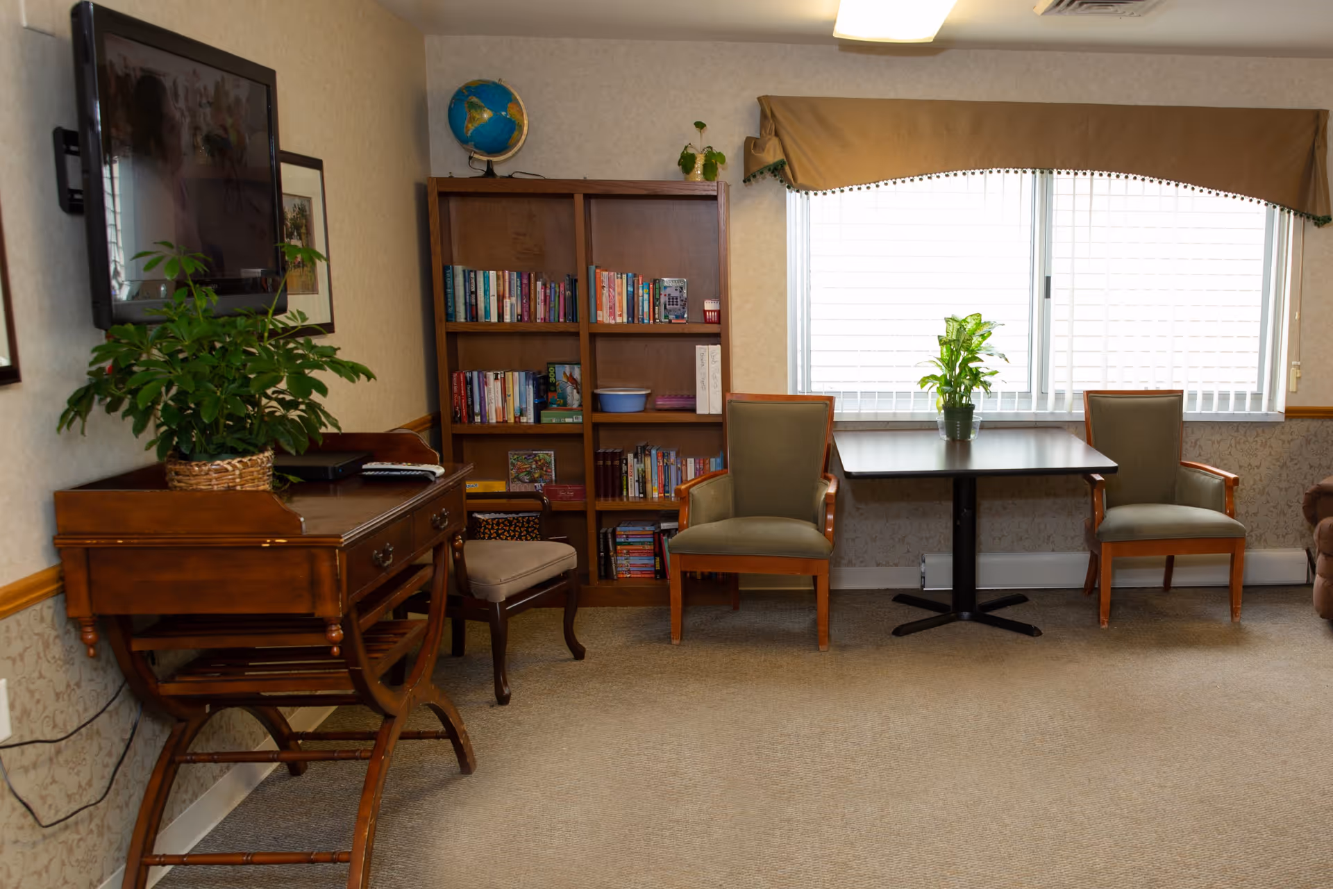 A cozy interior room with a wooden desk holding a potted plant and a TV mounted on the wall above it. Next to the desk is a wooden bookshelf filled with books and a globe on top. In front of a large window with a brown valance, there is a small table with a potted plant and two upholstered chairs on either side. The room has beige carpet and wallpaper with a wooden chair rail.