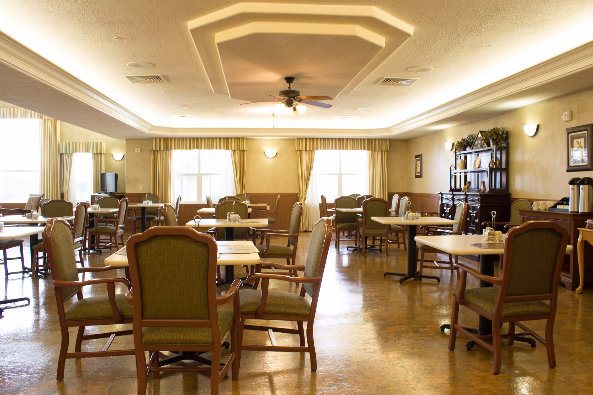A spacious dining room with multiple tables and green cushioned wooden chairs arranged neatly. The room has large windows with beige curtains allowing natural light to fill the space. There is a ceiling fan with lights in the center of the ceiling, and a wooden cabinet with decorative items against one wall. The floor has a polished finish and the overall ambiance is warm and inviting.