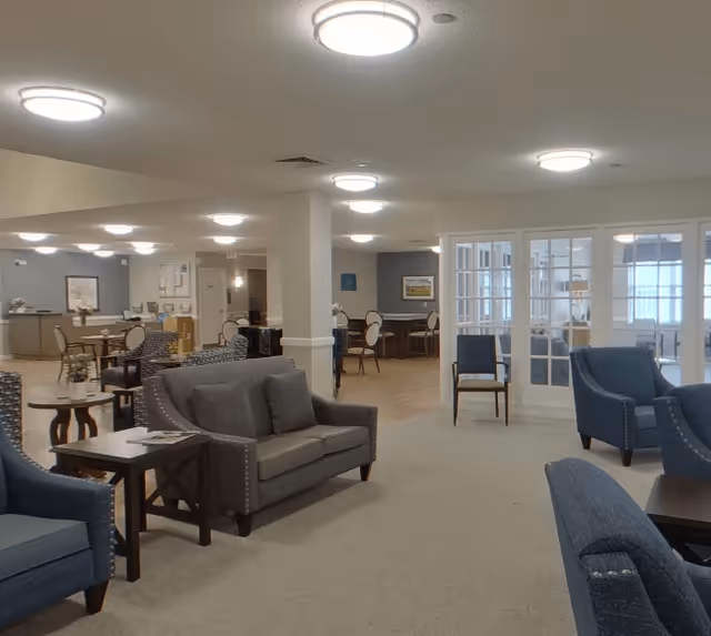 A spacious senior living facility common area with multiple seating arrangements including gray and blue upholstered chairs and sofas. The room has beige carpeting, white walls, and ceiling lights. In the background, there are dining tables and chairs, a reception or service desk, and a glass partition separating another room.