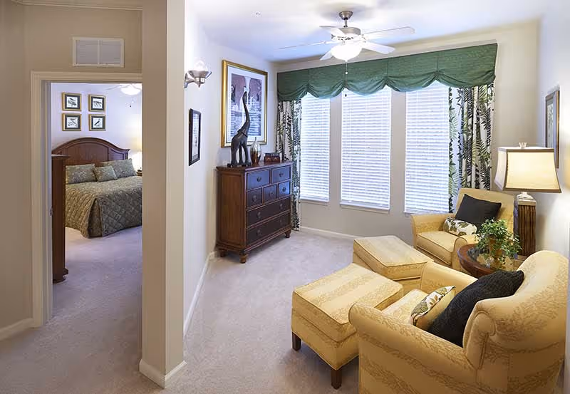 A cozy living room area with two beige armchairs, matching ottomans, a round wooden side table with a plant, and a floor lamp. The room has three large windows with white blinds and green valances with patterned curtains. A wooden dresser with decorative items and framed artwork is against the wall. To the left, an open doorway leads to a bedroom with a bed covered in a green quilt and framed pictures above the headboard.