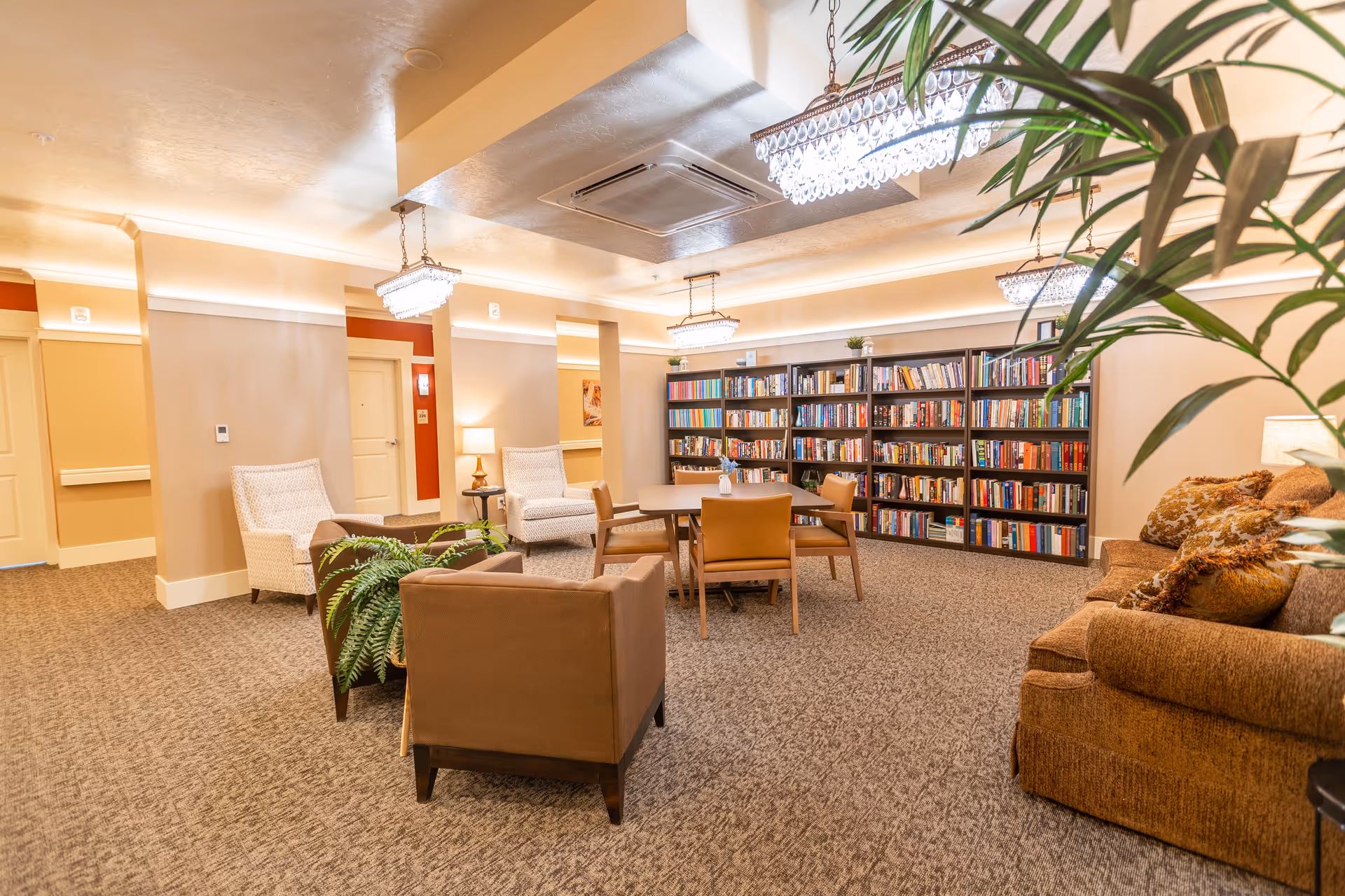 Cozy common room with bookshelves, seating, a central table and chandeliers in an assisted living facility.