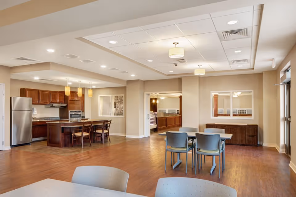 A spacious, well-lit room featuring a modern kitchen area with wooden cabinets, a stainless steel refrigerator, and an island with three chairs. Adjacent to the kitchen is a dining area with tables and chairs on a wooden floor, beige walls, and ceiling lights. There is a window and a door leading to another room in the background.