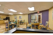 Interior view of a dining area in a senior living facility with several wooden tables and chairs arranged neatly. The walls are painted in light yellow and purple, with framed artwork hanging. The floor has a wood pattern, and there is a kitchen area visible in the background with cabinets and appliances.