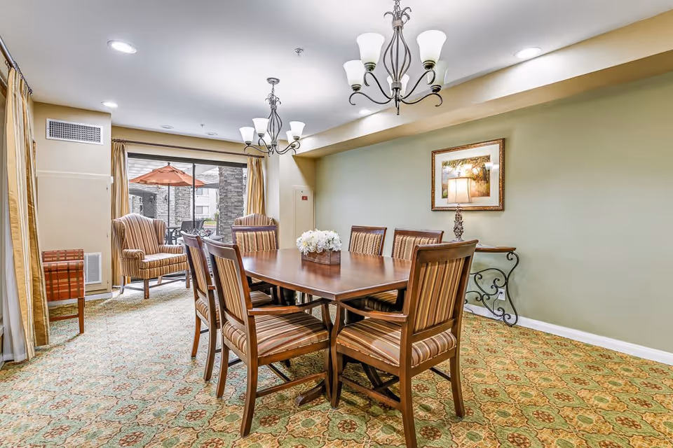 A well-lit dining room with a wooden dining table surrounded by eight striped upholstered chairs. Two chandeliers hang from the ceiling above the table. In the background, there are two armchairs near a large window with beige curtains, and outside the window, a patio with an orange umbrella is visible. The room has a green patterned carpet, light green walls, and a framed picture with a lamp on a decorative side table.