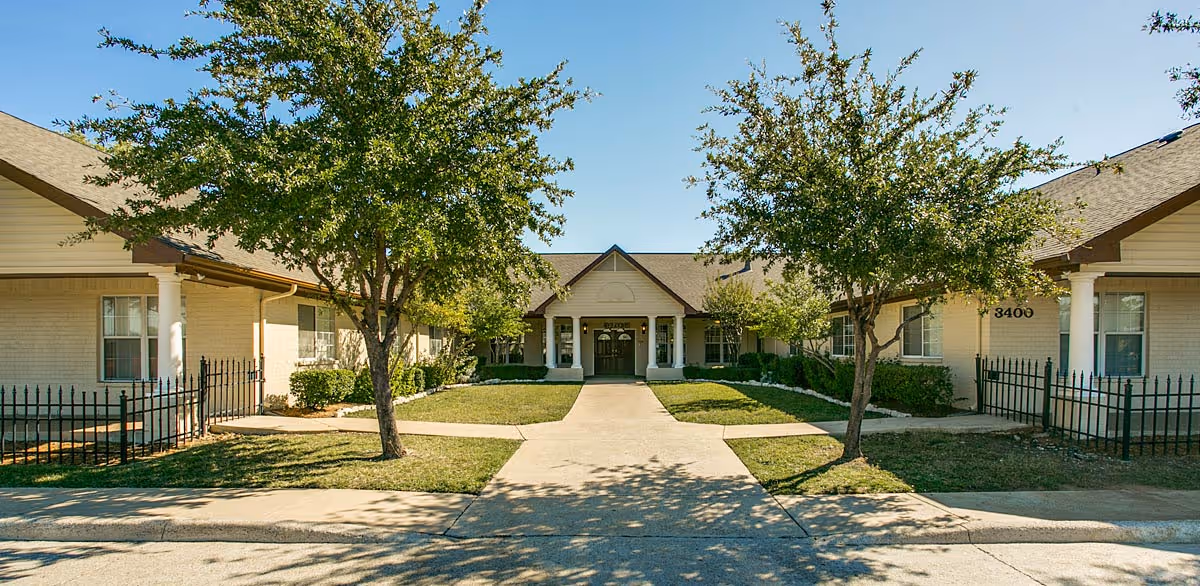 Senior living facility with a central entrance, beige-brick building, walkway, and trees in front yard.