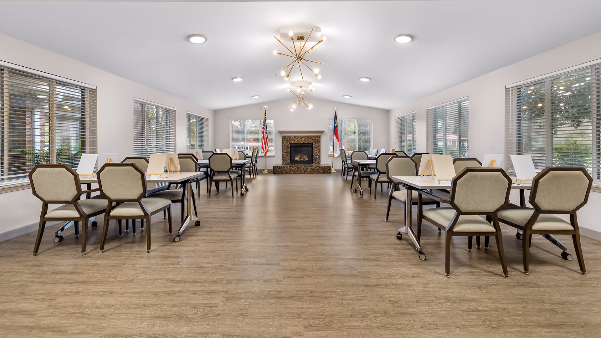 A spacious dining room with multiple tables and chairs arranged neatly on both sides. The room has large windows with blinds allowing natural light to enter. At the far end, there is a fireplace flanked by two flags. Modern light fixtures hang from the ceiling, and the floor has a wood-like finish.