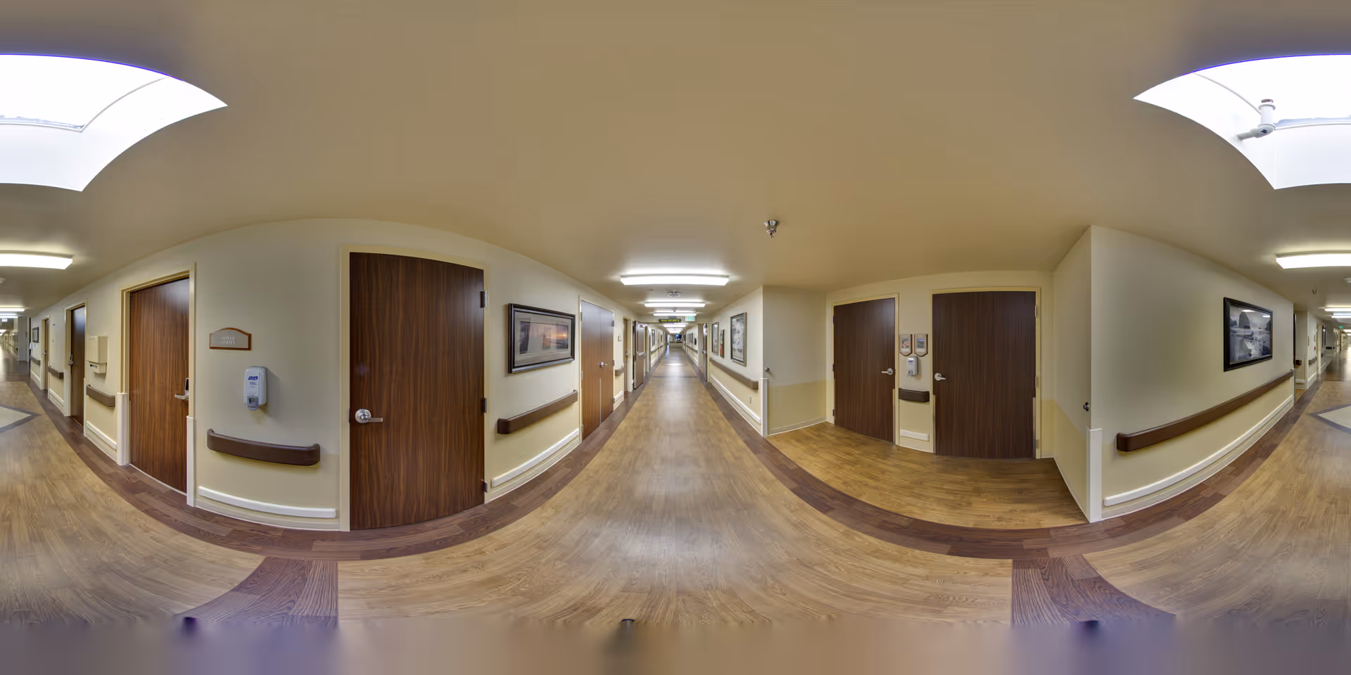 A long, well-lit hallway in a senior living facility with wooden doors on both sides, handrails along the walls, framed pictures hanging, and a wood-patterned floor. The ceiling has recessed lighting and skylights.