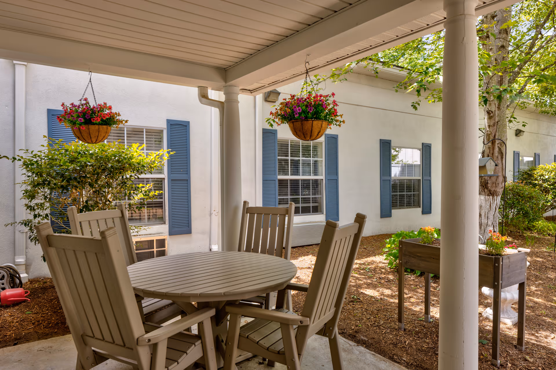 Covered outdoor patio area with a round table and four wooden chairs. Two hanging baskets with colorful flowers are suspended from the ceiling. The patio overlooks a garden area with plants, a tree, and a raised planter box. The building exterior has white walls and blue window shutters.