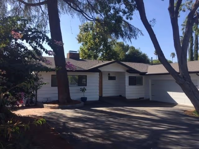 White, single-story building exterior with trees, driveway, and a shaded entrance.