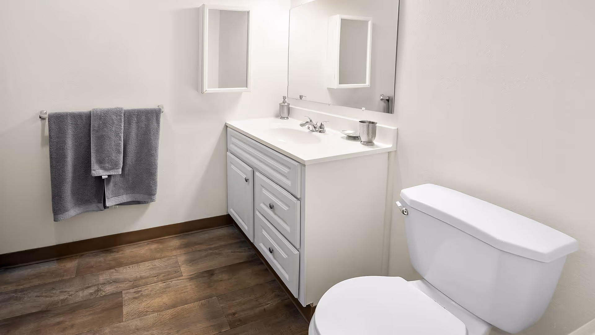 A clean bathroom with a white vanity cabinet featuring a sink and faucet, a large mirror above it, a white toilet, and two gray towels hanging on a towel rack on the wall. The floor has wood-like tiles and the walls are painted white.